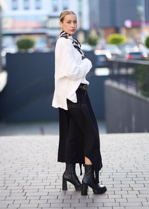 Czarna spódnica dzianinowa o asymetrycznym dole i elastycznym pasie z ozdobnym paskiem. Model pokazany w stylizacji z jasnym swetrem i czarnymi botkami na obcasie.