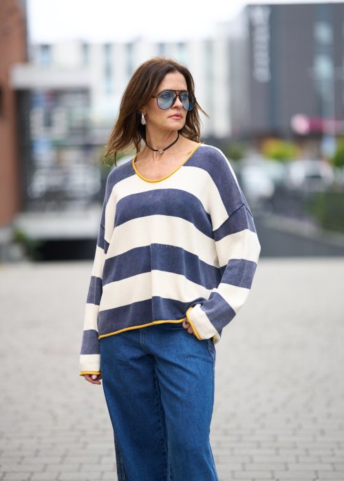 Kobieta w oversize swetrze w szerokie granatowo-kremowe pasy z żółtą lamówką, luźna stylizacja z jeansami, moda damska casual