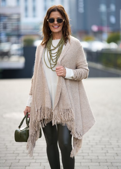 Beżowa narzutka z miękkiej dzianiny, luźny krój oversize i długie frędzle, styl boho w modzie damskiej
