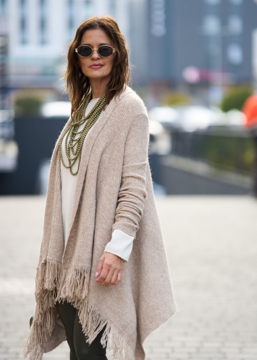Beżowa narzutka z miękkiej dzianiny, luźny krój oversize i długie frędzle, styl boho w modzie damskiej