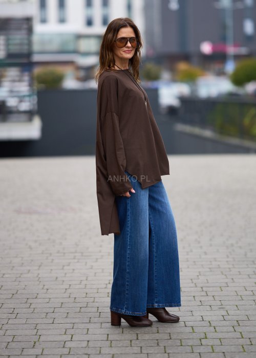 Czekoladowa bluzka oversize z dłuższym tyłem i długim rękawem, zestawiona z niebieskimi jeansami w swobodnej, miejskiej stylizacji.