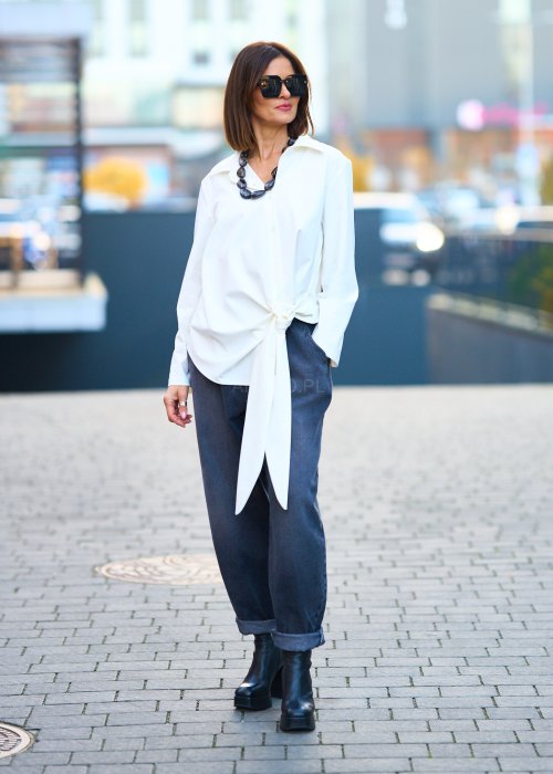 Biała koszula damska z asymetrycznym wiązaniem i klasycznym kołnierzem. Stylizacja z ciemnymi spodniami i biżuterią w eleganckim, miejskim stylu.