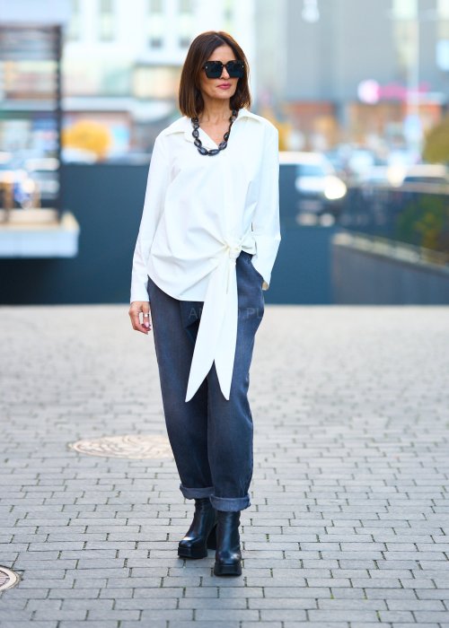 Biała koszula damska z asymetrycznym wiązaniem i klasycznym kołnierzem. Stylizacja z ciemnymi spodniami i biżuterią w eleganckim, miejskim stylu.