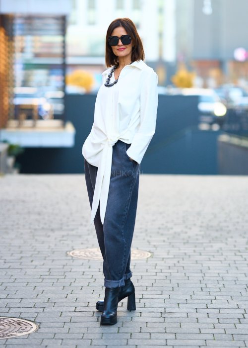 Biała koszula damska z asymetrycznym wiązaniem i klasycznym kołnierzem. Stylizacja z ciemnymi spodniami i biżuterią w eleganckim, miejskim stylu.