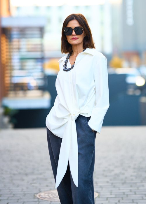 Biała koszula damska z asymetrycznym wiązaniem i klasycznym kołnierzem. Stylizacja z ciemnymi spodniami i biżuterią w eleganckim, miejskim stylu.
