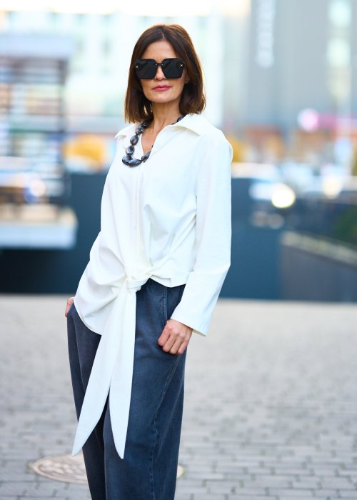 Biała koszula damska z asymetrycznym wiązaniem i klasycznym kołnierzem. Stylizacja z ciemnymi spodniami i biżuterią w eleganckim, miejskim stylu.