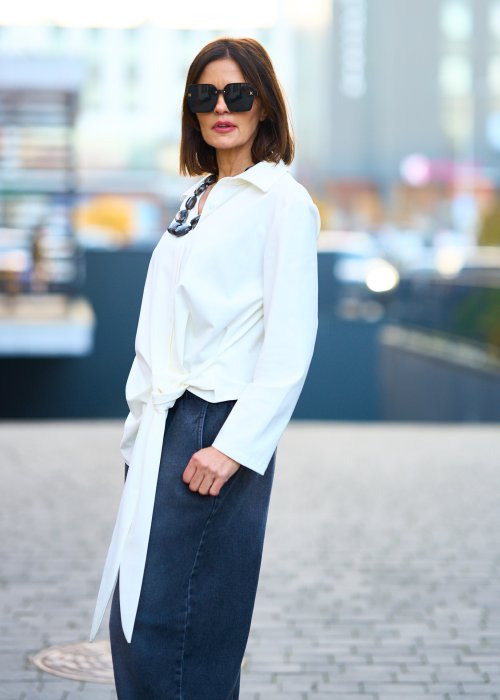 Biała koszula damska z asymetrycznym wiązaniem i klasycznym kołnierzem. Stylizacja z ciemnymi spodniami i biżuterią w eleganckim, miejskim stylu.