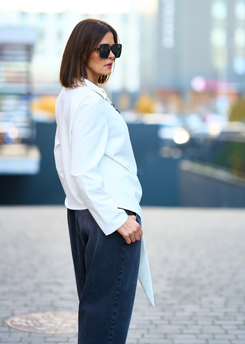 Biała koszula damska z asymetrycznym wiązaniem i klasycznym kołnierzem. Stylizacja z ciemnymi spodniami i biżuterią w eleganckim, miejskim stylu.