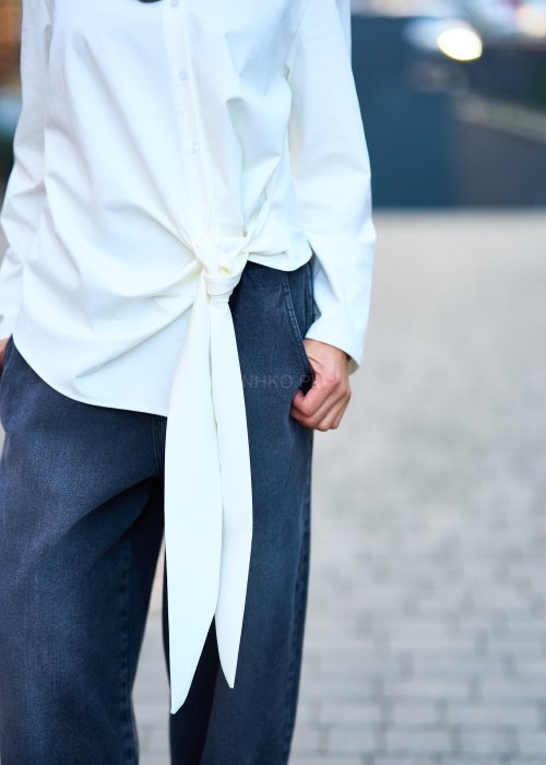 Biała koszula damska z asymetrycznym wiązaniem i klasycznym kołnierzem. Stylizacja z ciemnymi spodniami i biżuterią w eleganckim, miejskim stylu.