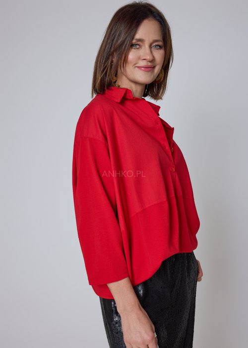 Czerwona koszula damska oversize z kołnierzykiem i przeszyciem poniżej biustu, zestawiona z czarnymi spodniami z połyskiem w eleganckiej stylizacji.