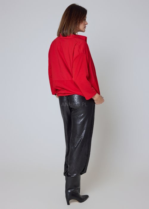 Czerwona koszula damska oversize z kołnierzykiem i przeszyciem poniżej biustu, zestawiona z czarnymi spodniami z połyskiem w eleganckiej stylizacji.