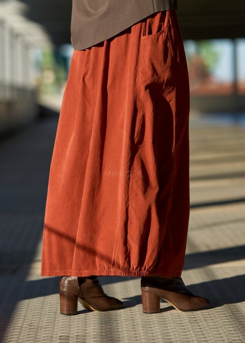 Ceglana spódnica maxi z elastyczną talią i kieszeniami, o luźnym kroju, zestawiona z brązową marynarką i skórzanymi botkami na obcasie w eleganckiej, miejskiej stylizacji.