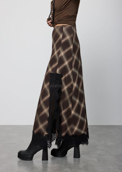 Długa damska spódnica w brązową kratę, o prostym, lekko rozszerzanym kroju; wykończona u dołu czarną koronką, noszona z czarnymi botkami na platformie.