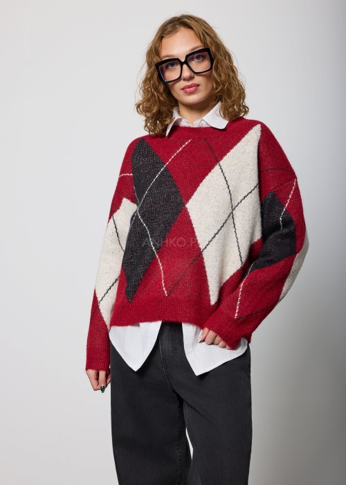 Damski sweter w romby w odcieniach czerwieni, czerni i ecru, o luźnym kroju i długich rękawach, noszony na białej koszuli z kołnierzykiem, zestawiony z czarnymi jeansami.