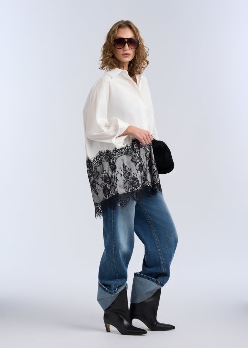 Biała koszula oversize z czarną, dekoracyjną koronką u dołu, długi rękaw i klasyczne zapięcie na guziki, stylizacja z jeansami.