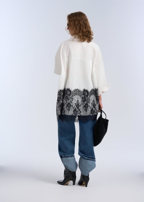 Biała koszula oversize z czarną, dekoracyjną koronką u dołu, długi rękaw i klasyczne zapięcie na guziki, stylizacja z jeansami.