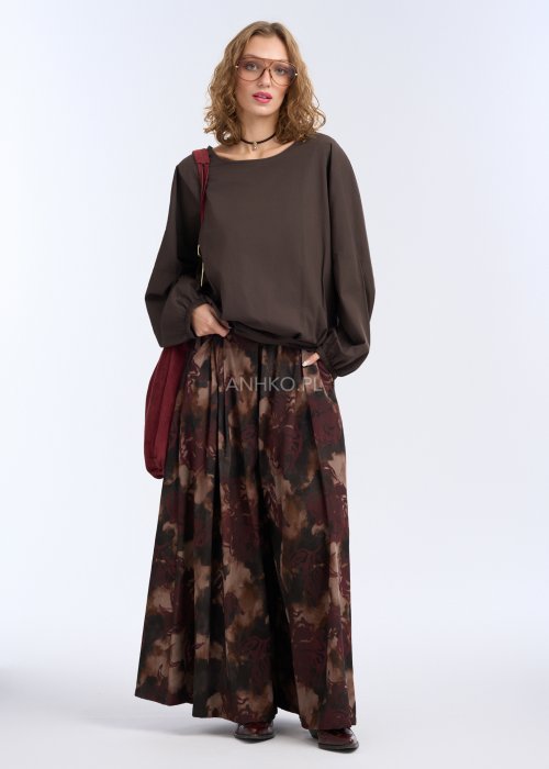 Modelka w luźnej, brązowej bluzce z długim rękawem i obniżoną linią ramion; bluzka o prostym kroju z delikatnym marszczeniem przy mankietach, zestawiona z szerokimi spodniami w abstrakcyjny wzór