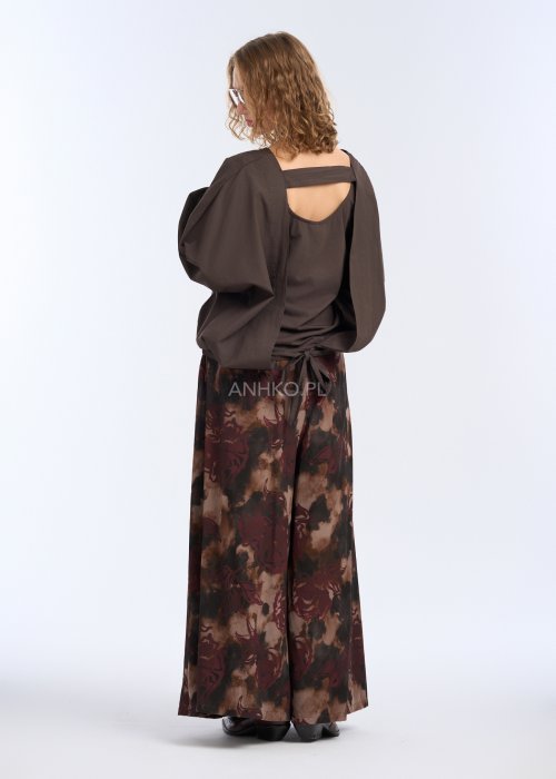 Modelka w luźnej, brązowej bluzce z długim rękawem i obniżoną linią ramion; bluzka o prostym kroju z delikatnym marszczeniem przy mankietach, zestawiona z szerokimi spodniami w abstrakcyjny wzór