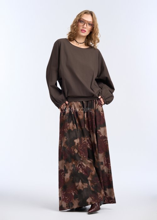 Modelka w luźnej, brązowej bluzce z długim rękawem i obniżoną linią ramion; bluzka o prostym kroju z delikatnym marszczeniem przy mankietach, zestawiona z szerokimi spodniami w abstrakcyjny wzór