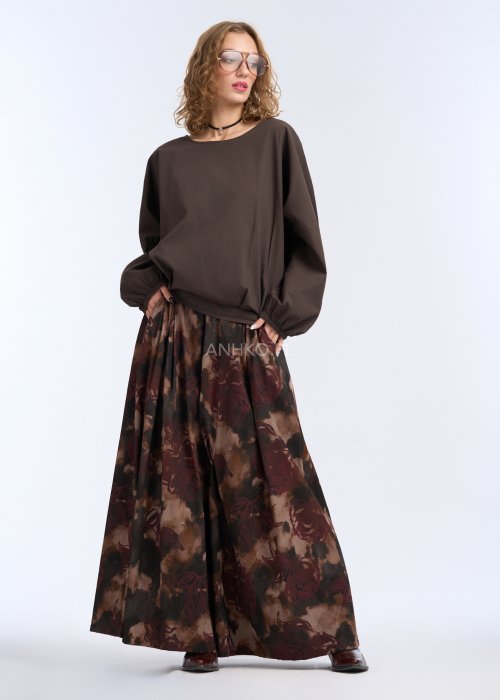 Modelka w luźnej, brązowej bluzce z długim rękawem i obniżoną linią ramion; bluzka o prostym kroju z delikatnym marszczeniem przy mankietach, zestawiona z szerokimi spodniami w abstrakcyjny wzór