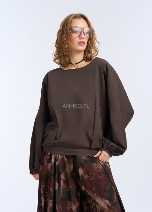Modelka w luźnej, brązowej bluzce z długim rękawem i obniżoną linią ramion; bluzka o prostym kroju z delikatnym marszczeniem przy mankietach, zestawiona z szerokimi spodniami w abstrakcyjny wzór