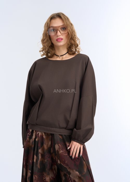 Modelka w luźnej, brązowej bluzce z długim rękawem i obniżoną linią ramion; bluzka o prostym kroju z delikatnym marszczeniem przy mankietach, zestawiona z szerokimi spodniami w abstrakcyjny wzór