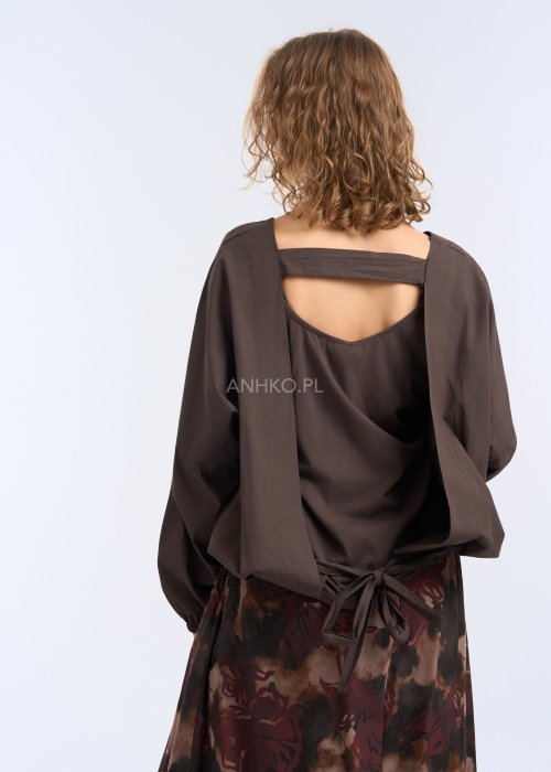 Modelka w luźnej, brązowej bluzce z długim rękawem i obniżoną linią ramion; bluzka o prostym kroju z delikatnym marszczeniem przy mankietach, zestawiona z szerokimi spodniami w abstrakcyjny wzór