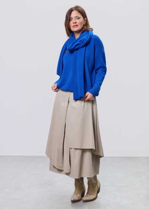 Kobaltowy sweter damski z miękkiej dzianiny, zintegrowany z długim szalem luźno opadającym przy dekolcie; model o swobodnym kroju, długi rękaw, stylizowany na jasnym tle