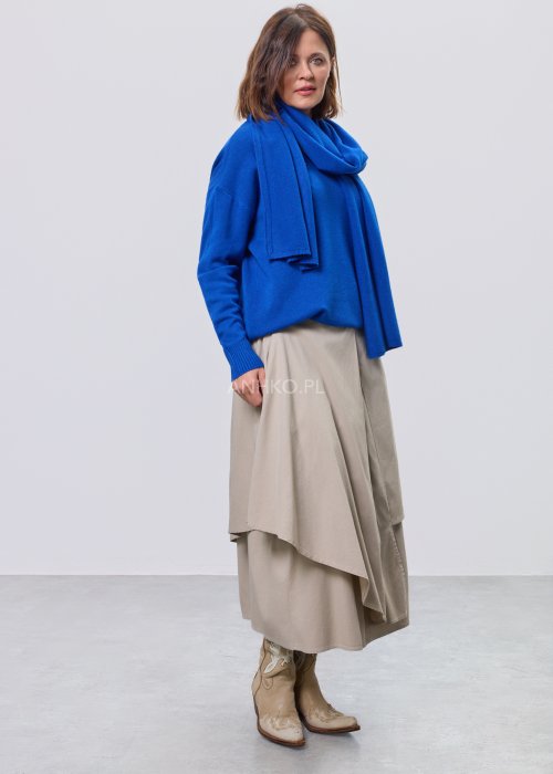 Kobaltowy sweter damski z miękkiej dzianiny, zintegrowany z długim szalem luźno opadającym przy dekolcie; model o swobodnym kroju, długi rękaw, stylizowany na jasnym tle