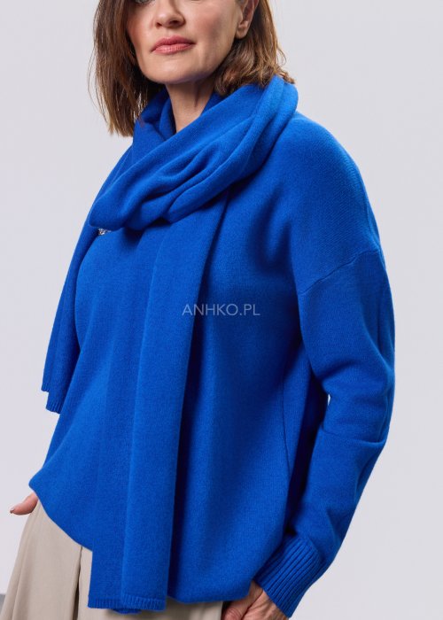 Kobaltowy sweter damski z miękkiej dzianiny, zintegrowany z długim szalem luźno opadającym przy dekolcie; model o swobodnym kroju, długi rękaw, stylizowany na jasnym tle