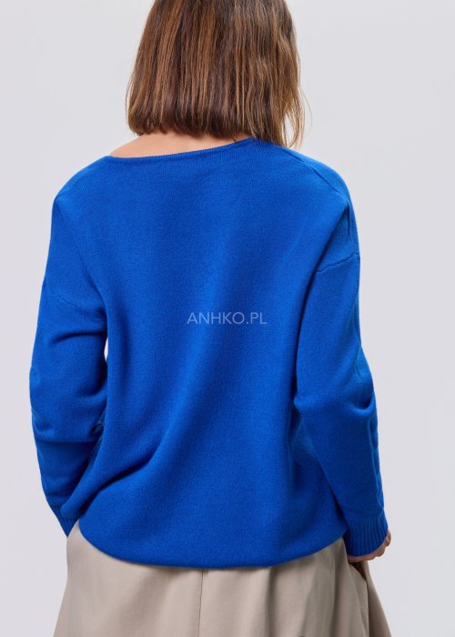 Kobaltowy sweter damski z miękkiej dzianiny, zintegrowany z długim szalem luźno opadającym przy dekolcie; model o swobodnym kroju, długi rękaw, stylizowany na jasnym tle