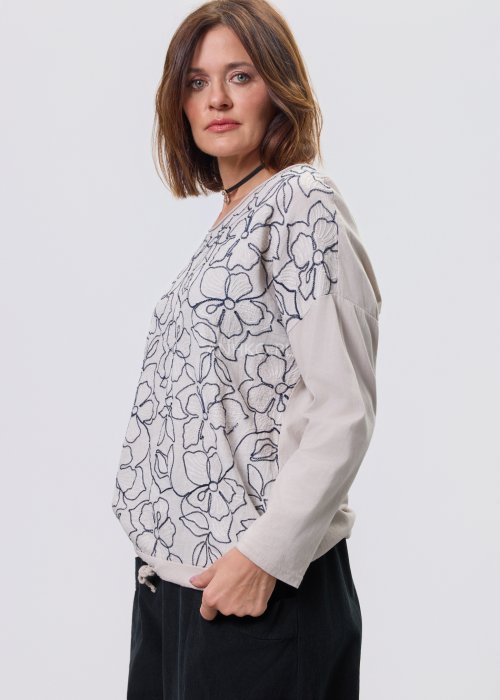 Jasna bluzka damska z długim rękawem, wykonana z lekkiej tkaniny, z dekoracyjnym haftem w kontrastowy kwiatowy wzór na przodzie; prosty, swobodny krój, okrągły dekolt, stylizacja na jasnym tle