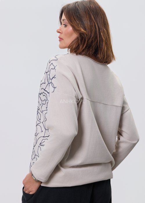 Jasna bluzka damska z długim rękawem, wykonana z lekkiej tkaniny, z dekoracyjnym haftem w kontrastowy kwiatowy wzór na przodzie; prosty, swobodny krój, okrągły dekolt, stylizacja na jasnym tle