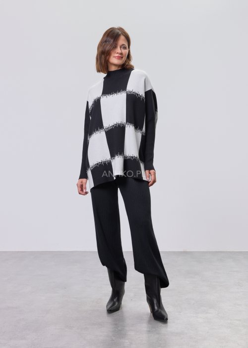 Czarno-biała damska tunika dzianinowa o luźnym kroju, z półgolfem i długimi rękawami, w geometryczny wzór pasów z delikatnym motywem graficznym, prezentowana na modelce na jasnym tle
