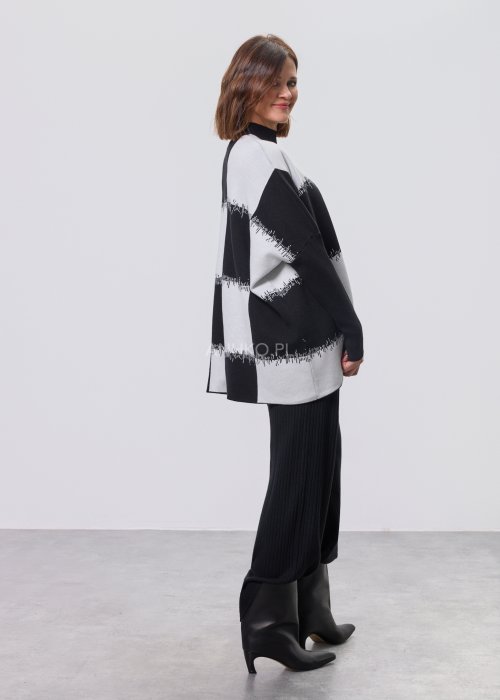 Czarno-biała damska tunika dzianinowa o luźnym kroju, z półgolfem i długimi rękawami, w geometryczny wzór pasów z delikatnym motywem graficznym, prezentowana na modelce na jasnym tle