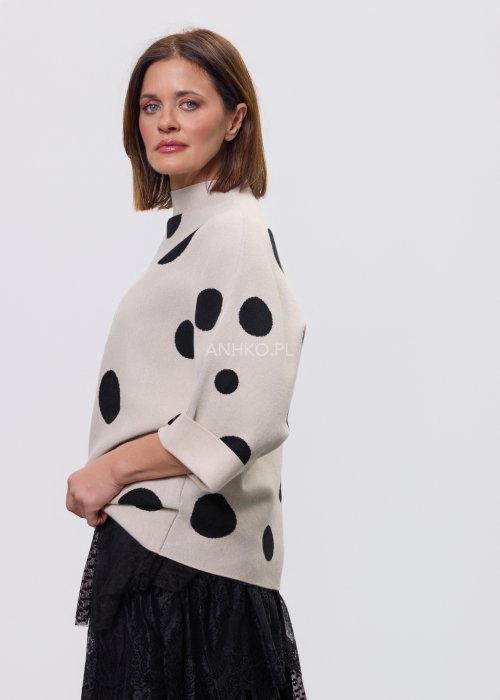 Jasny, beżowy damski sweter o luźnym kroju z półgolfem i rękawami 3/4, ozdobiony kontrastowym wzorem czarnych kropek, prezentowany na modelce na jasnym tle