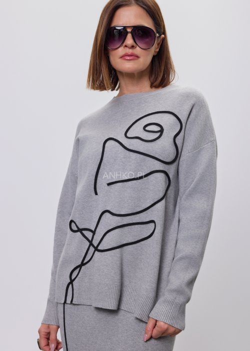 Modelka w szarym, dzianinowym komplecie dwuczęściowym: luźny sweter z długim rękawem i czarnym abstrakcyjnym nadrukiem z przodu oraz dopasowana spódnica midi w tym samym kolorze