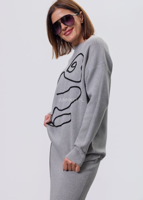 Modelka w szarym, dzianinowym komplecie dwuczęściowym: luźny sweter z długim rękawem i czarnym abstrakcyjnym nadrukiem z przodu oraz dopasowana spódnica midi w tym samym kolorze