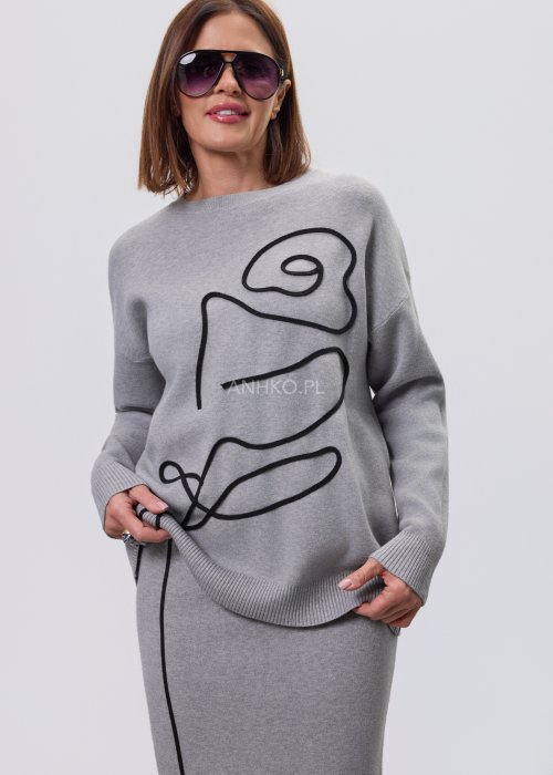 Modelka w szarym, dzianinowym komplecie dwuczęściowym: luźny sweter z długim rękawem i czarnym abstrakcyjnym nadrukiem z przodu oraz dopasowana spódnica midi w tym samym kolorze