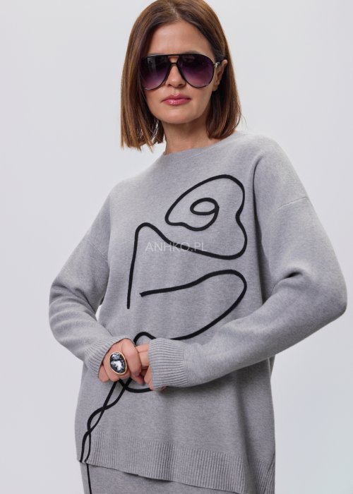 Modelka w szarym, dzianinowym komplecie dwuczęściowym: luźny sweter z długim rękawem i czarnym abstrakcyjnym nadrukiem z przodu oraz dopasowana spódnica midi w tym samym kolorze