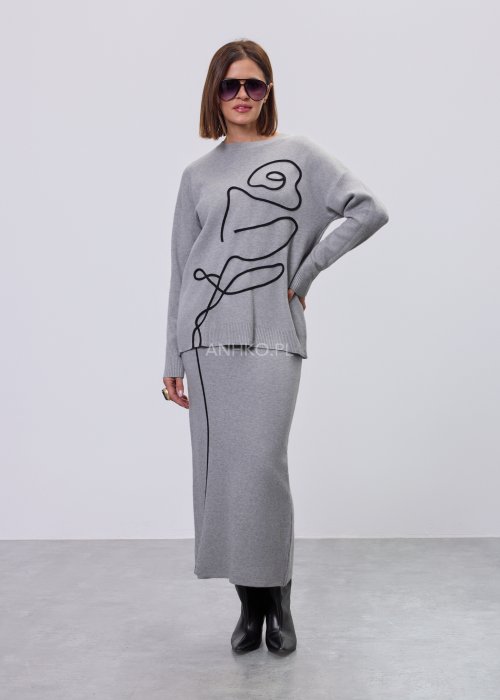 Modelka w szarym, dzianinowym komplecie dwuczęściowym: luźny sweter z długim rękawem i czarnym abstrakcyjnym nadrukiem z przodu oraz dopasowana spódnica midi w tym samym kolorze