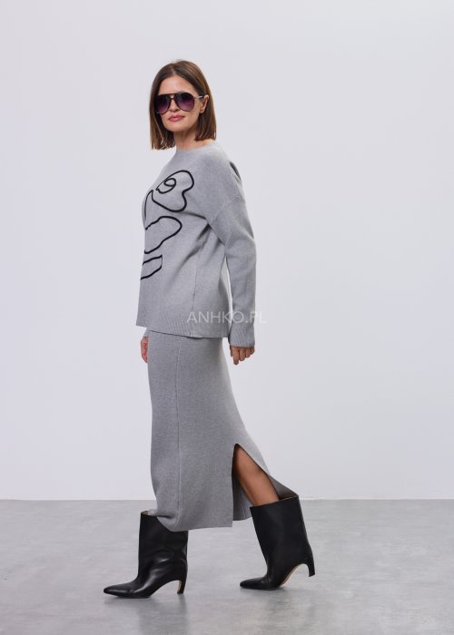 Modelka w szarym, dzianinowym komplecie dwuczęściowym: luźny sweter z długim rękawem i czarnym abstrakcyjnym nadrukiem z przodu oraz dopasowana spódnica midi w tym samym kolorze