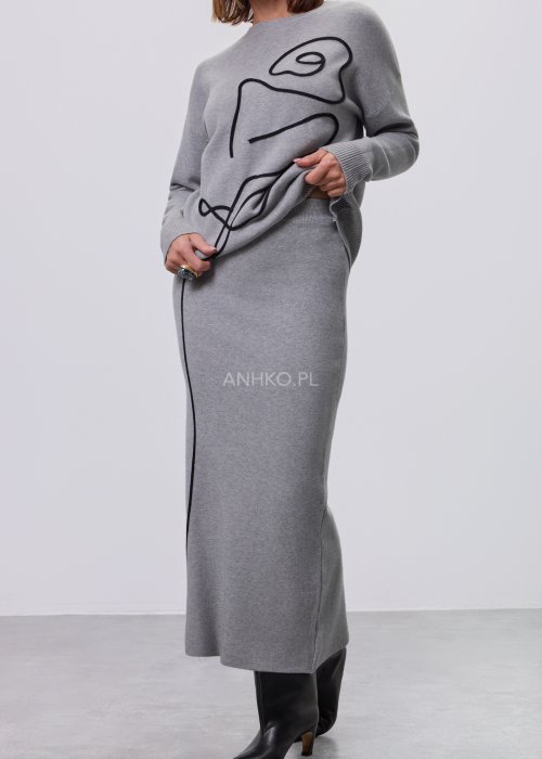 Modelka w szarym, dzianinowym komplecie dwuczęściowym: luźny sweter z długim rękawem i czarnym abstrakcyjnym nadrukiem z przodu oraz dopasowana spódnica midi w tym samym kolorze