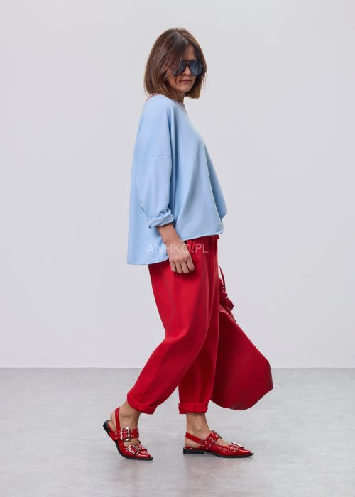 Niebieska bluza damska oversize w odcieniu baby blue z asymetrycznym dołem i kontrastowym dekoltem. Stylizacja z czerwonymi spodniami podkreśla nowoczesny charakter modelu.