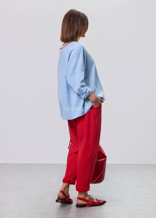 Niebieska bluza damska oversize w odcieniu baby blue z asymetrycznym dołem i kontrastowym dekoltem. Stylizacja z czerwonymi spodniami podkreśla nowoczesny charakter modelu.