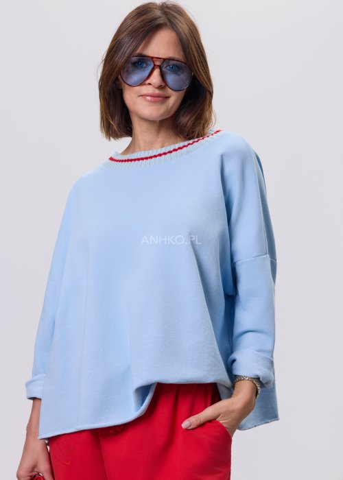 Niebieska bluza damska oversize w odcieniu baby blue z asymetrycznym dołem i kontrastowym dekoltem. Stylizacja z czerwonymi spodniami podkreśla nowoczesny charakter modelu.
