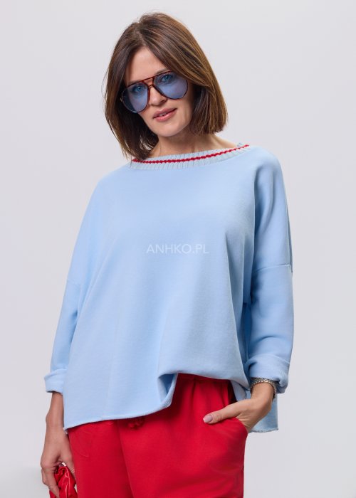 Niebieska bluza damska oversize w odcieniu baby blue z asymetrycznym dołem i kontrastowym dekoltem. Stylizacja z czerwonymi spodniami podkreśla nowoczesny charakter modelu.