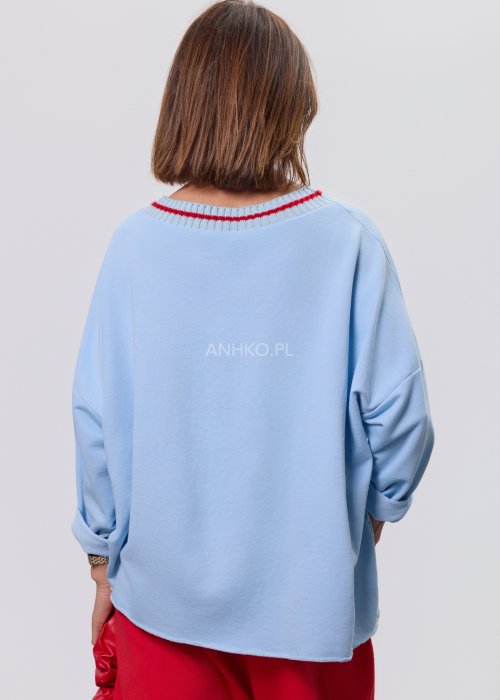 Niebieska bluza damska oversize w odcieniu baby blue z asymetrycznym dołem i kontrastowym dekoltem. Stylizacja z czerwonymi spodniami podkreśla nowoczesny charakter modelu.