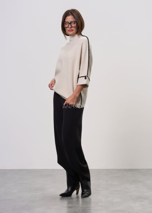 Beżowa damska bluzka o luźnym kroju, z półgolfem i rękawami 3/4, wykończona kontrastową czarną lamówką wzdłuż ramion, rękawów i boków oraz ozdobnymi guzikami przy mankietach.