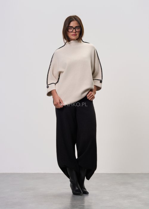 Beżowa damska bluzka o luźnym kroju, z półgolfem i rękawami 3/4, wykończona kontrastową czarną lamówką wzdłuż ramion, rękawów i boków oraz ozdobnymi guzikami przy mankietach.